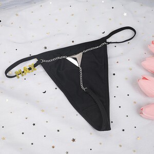 Könnte beinhalten: Schwarzer String-Tanga mit silberner Kette und dem Wort "MAX" in goldfarbenen Glitzerbuchstaben. Der String besteht aus einem weichen Material und hat ein minimalistisches Design.