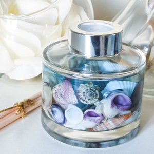 May include: A clear glass diffuser bottle filled with blue liquid and various seashells. The bottle has a silver lid and is surrounded by beige reeds.