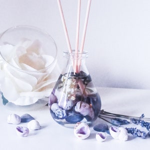 May include: A clear glass diffuser bottle with a pearlescent finish filled with water, seashells, and lavender. The bottle has a set of wooden diffuser sticks.