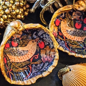 May include: Hand-painted seashell ornaments with a bird and floral design. The interior of the shells features a dark background with red apples and a bird. The edges are trimmed in gold, and a gold string is attached for hanging.