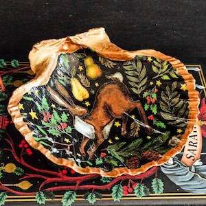May include: A decorative shell dish with a painted interior. The design features a brown hare, pears, and foliage on a black background. The shell's edge is gold. A unique piece of home decor, perfect for displaying small items or as a decorative accent.