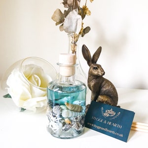 May include: A decorative glass bottle with blue liquid and seashells, sealed with a wooden stopper. A bronze hare ornament and a business card with the text "HYGGE & HEARTH" are also visible.