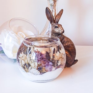 May include: A clear glass vase filled with dried flowers and a white sand base. The vase is sitting on a table next to a brown rabbit figurine.