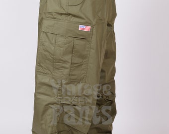 May include: A pair of olive green cargo pants with a drawstring waist and a small American flag patch on the left leg. The pants are loose fitting and have multiple pockets.