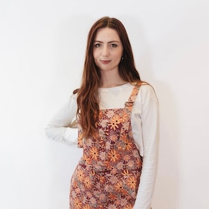 May include: Overalls with a floral pattern in shades of burgundy, orange, and yellow. The overalls have adjustable straps and a front pocket. A long-sleeved, off-white shirt is worn underneath.