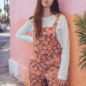 May include: Overalls with a floral pattern in shades of orange, yellow, and burgundy. The overalls are worn over a white long-sleeve shirt. The model is leaning against a pink and orange wall.