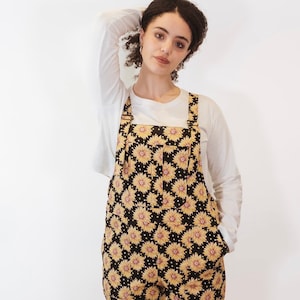 May include: Black overalls patterned with yellow sunflowers. The dungarees have adjustable straps and pockets. A white long-sleeved top is worn underneath. The overalls are made of a soft fabric.
