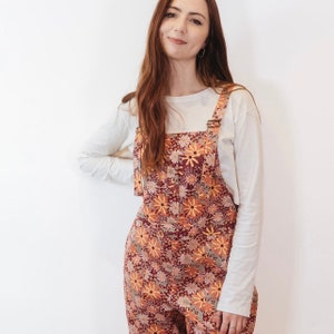 May include: Floral print overalls in shades of burgundy, orange, and yellow. The overalls feature a daisy pattern and adjustable straps. A long-sleeved white shirt is worn underneath. The outfit is suitable for casual wear.