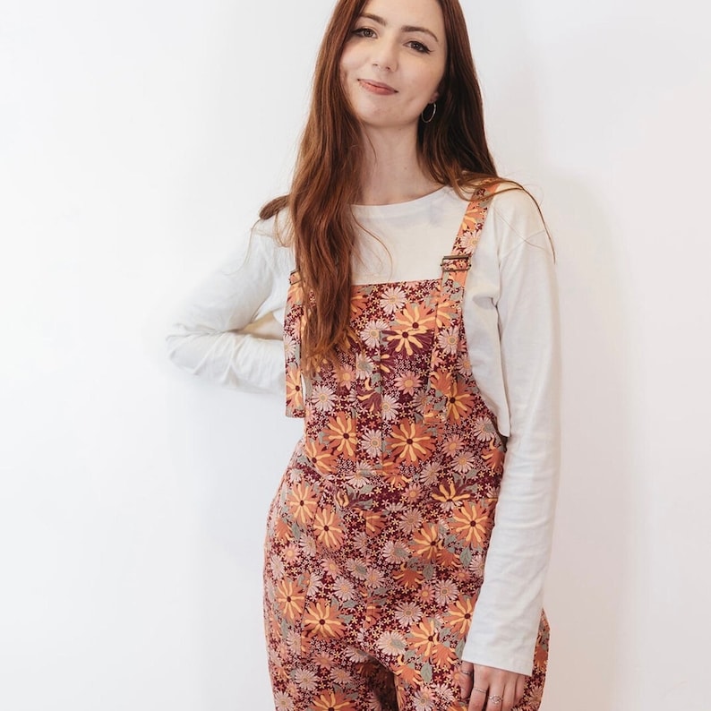 May include: Floral print overalls in shades of burgundy, orange, and yellow. The overalls feature a daisy pattern and adjustable straps. A long-sleeved white shirt is worn underneath. The outfit is suitable for casual wear.