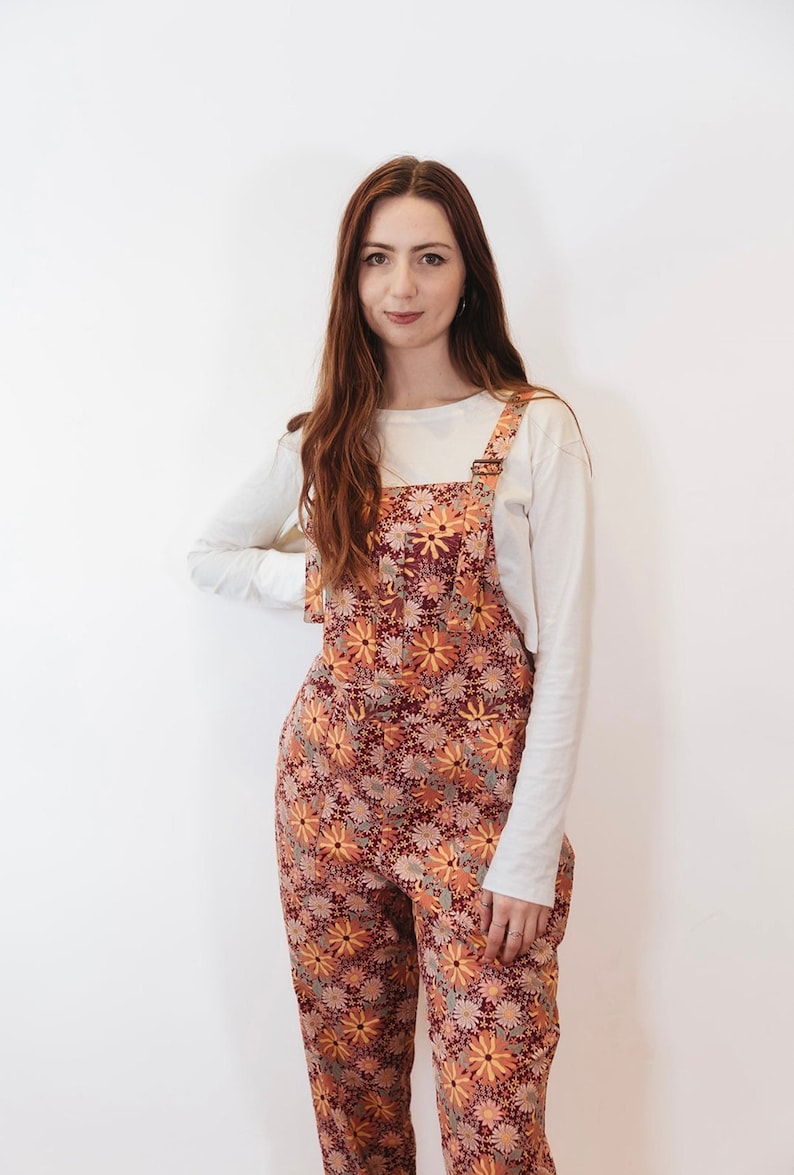 May include: Overalls with a floral pattern in shades of burgundy, orange, and yellow. The overalls have adjustable straps and a front pocket. A long-sleeved, off-white shirt is worn underneath.
