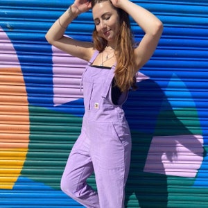 May include: A woman wearing a lavender colored overall jumpsuit with a single pocket on the front. The overall has thin straps and is made of a soft, textured fabric. The woman is standing in front of a colorful wall.