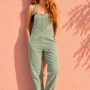 May include: Light green corduroy overalls with adjustable straps and a front pocket. The overalls have a relaxed fit and are cuffed at the ankles. A small logo patch is visible on the chest pocket. The model is standing in front of a pink wall.