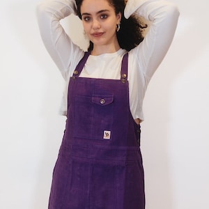 May include: Purple corduroy dungarees with a small patch on the front pocket. The dungarees have adjustable straps and a button closure.