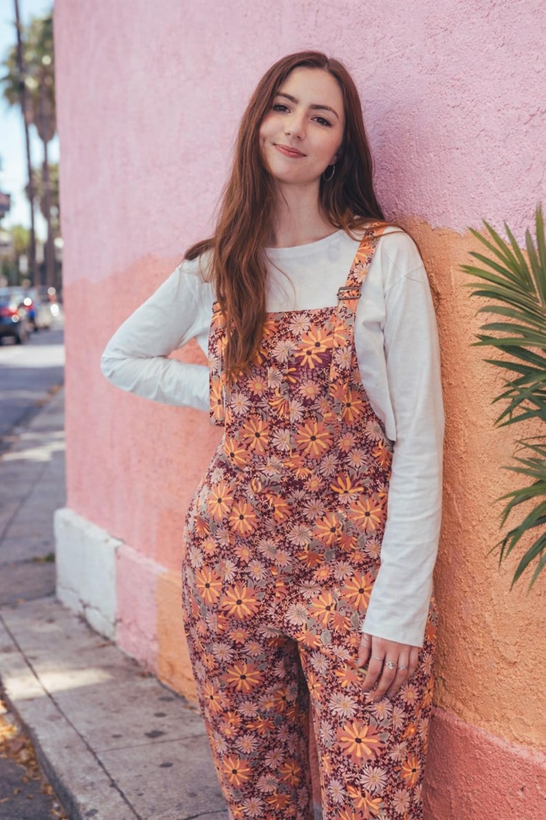 May include: Overalls with a floral pattern in shades of orange, yellow, and burgundy. The overalls are worn over a white long-sleeve shirt. The model is leaning against a pink and orange wall.