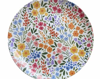 May include: A white round plate with a floral pattern. The pattern features a variety of colorful flowers, including pink, orange, yellow, blue, and purple. The flowers are arranged in a random pattern on the plate.