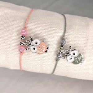Könnte beinhalten: Zwei Freundschaftsarmbänder mit Eulenanhängern. Ein Armband hat rosa Perlen und einen pfirsichfarbenen Eulenanhänger. Das andere Armband hat graue Perlen und einen grauen Eulenanhänger.