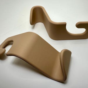 May include: Two tan-colored, curved plastic car seat hooks. Each hook has a rounded, hook-shaped end and a curved body. The hooks are designed to attach to a car seat and provide a place to hang items.