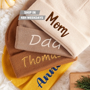 May include: Four knitted beanies in different colours with embroidered names: Mom, Dad, Thomas, and Anna. The beanies are stacked on top of each other.