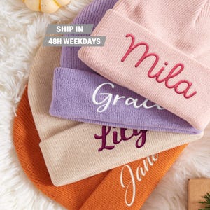 May include: Four knitted beanies in different colors with embroidered names: Mila, Grace, Lily, and Jane. The beanies are stacked on top of each other.