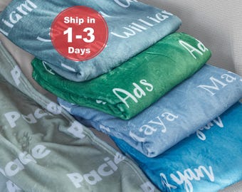 May include: A stack of four folded plush blankets in shades of teal, green, light blue, and sage. Each blanket features white text printed with various names including William, Ads, Maya, and Ryan, displayed in a repeating script font pattern.