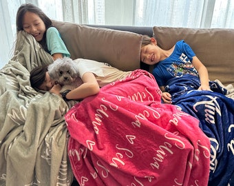 May include: Three children are relaxing on a couch with a small white dog. The children are covered in blankets with personalized text. The blanket on the left is gray, the middle blanket is pink with white text, and the blanket on the right is blue with white text.