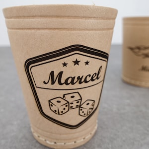 Könnte beinhalten: Zwei braune Pappbecher mit schwarzen Grafiken. Der vordere Becher zeigt ein Schilddesign mit dem Namen "Marcel" und Würfeln. Der zweite Becher hat ein Design und den Namen "Michael". Würfel sind im Hintergrund sichtbar. Die Becher sind für Spiele oder Veranstaltungen.