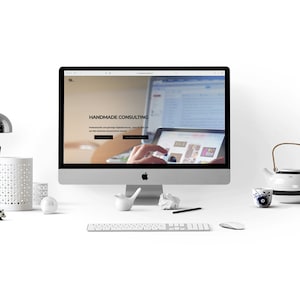 Könnte beinhalten: Ein weißer Desktop-Computer mit einer geöffneten Website auf dem Bildschirm. Die Website ist für ein Unternehmen namens "Handmade Consulting". Der Computer steht auf einem weißen Schreibtisch mit einer Lampe, einem Stifthalter, einer weißen Teekanne, einer Tasse und Kopfhörern.