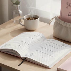 Peut inclure: Un journal ouvert avec des pages lignées et un signet en ruban noir est posé sur une surface en bois clair. Une tasse de thé fumante est à proximité, avec un petit vase et un journal dans un sac à cordon en arrière-plan. Le journal est ouvert.