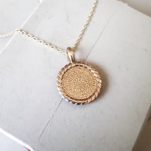 Op de afbeelding: Een gouden ketting met een ronde hanger met een cirkelvormig ontwerp met ingewikkelde details en een touwachtige rand.