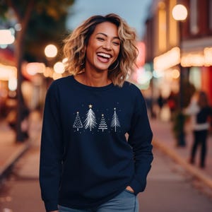 Könnte beinhalten: Marineblaues Sweatshirt mit einem weißen und goldenen Weihnachtsbaum-Design. Die Grafik zeigt vier stilisierte Bäume mit Sternakzenten. Das langärmlige Oberteil ist aus weichem Material und für die Weihnachtszeit geeignet.