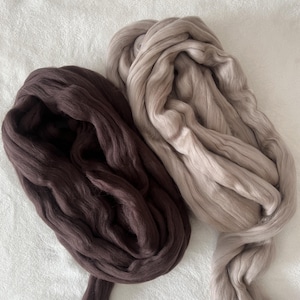 Könnte beinhalten: Zwei Stränge superklobiger Merinowolle in Braun- und Beigetönen. Die Wolle ist weich und flauschig, perfekt zum Armstricken oder für andere Handarbeiten. Die Stränge werden auf einer weißen Oberfläche präsentiert.