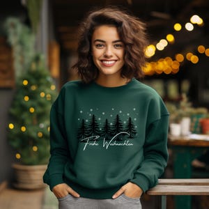 Könnte beinhalten: Ein dunkelgrünes Sweatshirt mit dem Text "Frohe Weihnachten" in weißer Schrift. Das Sweatshirt zeigt eine Grafik mit silhouettierten Kiefern und Schneeflocken. Die Person auf dem Bild lächelt und trägt eine graue Hose.