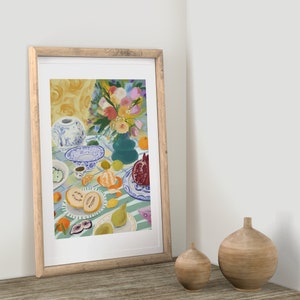 Pode incluir: Uma pintura emoldurada de uma natureza morta com um vaso de porcelana azul e branco cheio de flores coloridas, frutas e um prato com um padr&atilde;o azul e branco. A pintura est&aacute; colocada contra uma parede branca.