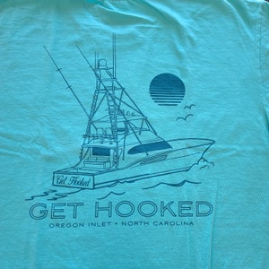 May include: A turquoise blue t-shirt with a graphic of a fishing boat and the text "Get Hooked Oregon Inlet North Carolina".