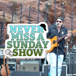 May include: A man with a black hat and sunglasses plays a guitar on stage. The stage is set up in front of a large rock formation. The text "NEVER MISS A SUNDAY SHOW" is visible in the foreground.