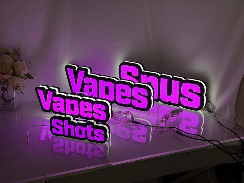 May include: Three illuminated signs with bright purple lettering and white outlines. The signs display the words Vapes, Snus, and Shots in a bold, blocky font. Each sign features a thin white power cord extending from the side, casting a glow.