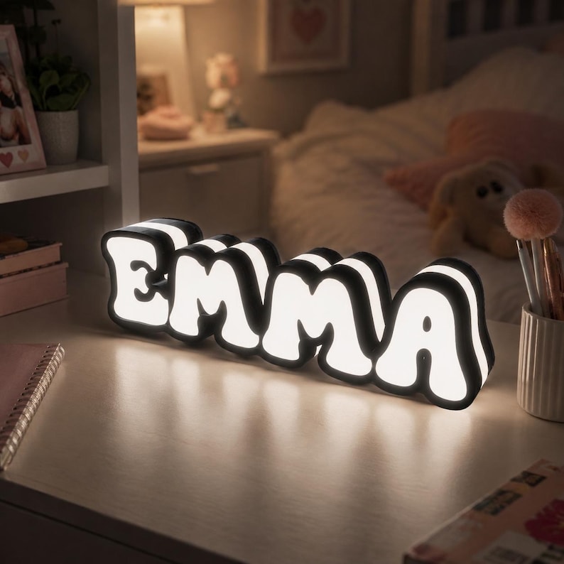 May include: A custom name sign featuring the letters EMMA in a bold, rounded font. The sign has a black outer frame and a bright white illuminated center, creating a glowing effect. It is a freestanding decorative light piece for a desk or shelf.