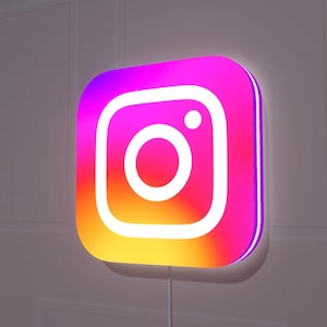 May include: A square wall-mounted sign with rounded corners featuring the Instagram logo. The sign displays a vibrant gradient of pink, purple, orange, and yellow colors with a bright white illuminated camera icon and dot centered on the front surface.