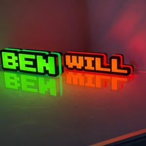 May include: Two custom 3D-printed signs with blocky, pixelated lettering. The left sign glows in bright green and displays the name BEN, while the right sign glows in vibrant orange and displays the name WILL. Both signs feature black borders.