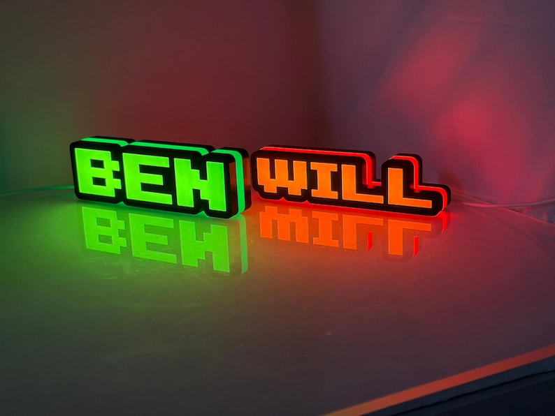 May include: Two custom 3D-printed signs with blocky, pixelated lettering. The left sign glows in bright green and displays the name BEN, while the right sign glows in vibrant orange and displays the name WILL. Both signs feature black borders.