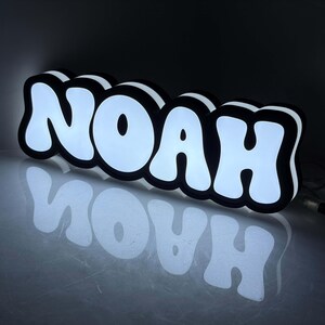 May include: A custom LED sign displaying the name NOAH in bold, white, bubble-style letters with a thick black outline. The sign emits a bright white glow from the front and sides, creating a reflective effect on the surface below it.