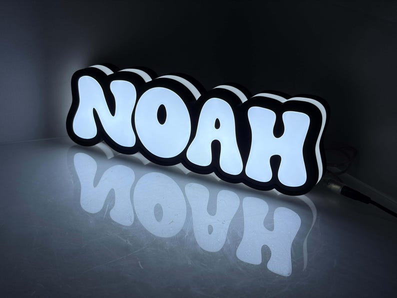May include: A custom LED sign displaying the name NOAH in bold, white, bubble-style letters with a thick black outline. The sign emits a bright white glow from the front and sides, creating a reflective effect on the surface below it.