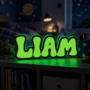May include: A custom-made, illuminated sign displaying the name LIAM in bold, rounded, lime green letters with a thick black border. The sign emits a bright glow, highlighting the typography against the dark, out-of-focus bedroom setting.