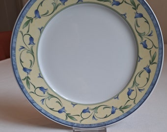 May include: A white ceramic dinner plate with a yellow and blue floral design. The plate has a blue rim and a yellow center with blue flowers and green leaves.