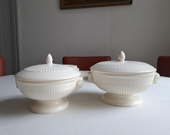 May include: Two off-white ceramic lidded tureens featuring a ribbed body design, a pedestal base, and decorative handles. Each tureen is topped with a matching lid that has a pointed finial. The surface of the tureens displays a subtle textured pattern.