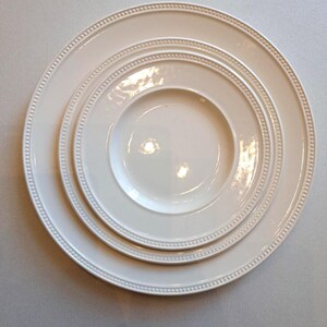 Puede incluir: Un juego de tres platos de cerámica de color blanco roto, apilados y de diferentes tamaños. Cada plato presenta un acabado liso y brillante con una textura de cuentas en relieve a lo largo del borde exterior, creando un diseño uniforme y limpio.