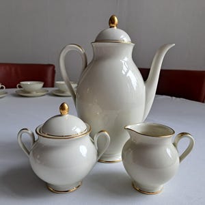 Puede incluir: Una cafetera de porcelana blanca con ribetes dorados, un azucarero y una jarra de crema a juego. El juego está sobre un mantel blanco.