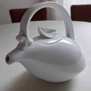 May include: A white ceramic teapot with a curved handle and a spout. The teapot has a smooth, glossy finish and a lid with a leaf-shaped detail. The handle is attached to the body with decorative swirls.