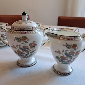 May include: A white porcelain tea set with a floral and bird design. The set includes a lidded sugar bowl, a creamer, and other pieces. The design features colorful flowers and a peacock.