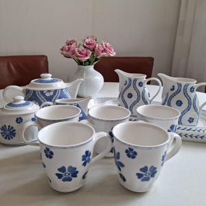 Op de afbeelding: Een keramische theeset met een witte basis en blauwe bloem- en touwpatronen. De set bevat een theepot met deksel, een suikerpot, twee kannetjes, diverse mokken en een ovaal dienblad, allemaal gerangschikt op een wit oppervlak.